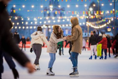 Uzun saçlı sarışın saçlı beyaz adam genç çift tr ve eğlenceli, aktif tarihi buz pateni Buz arenada akşam şehir meydanında Noel arifesinde kışın sakal ve güzel bir kadın var