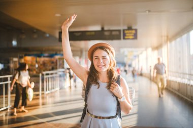 Tema Turizm ve seyahat. Genç güzel beyaz bir kadın elbise ve bir sırt çantası ile bir şapka. Uzun Koridor geçiş, bir zamanlama Yönetim Kurulu, bir el yükseltmek için bir kapı terminal istasyonu turistik mat.