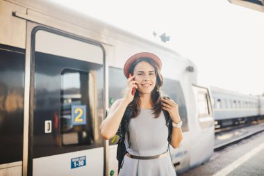 Tema ulaşım ve seyahat. Dişlek gülümseme ayakta portre Genç beyaz kadınla İstasyonu Tren arka plan teknolojisini kullanarak sırt çantası ile tren, telefon el elbise ve şapka yaz aylarında akıllı.