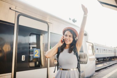 Tema ulaşım ve seyahat. Dişlek gülümseme ayakta portre Genç beyaz kadınla İstasyonu Tren arka plan teknolojisini kullanarak sırt çantası ile tren, telefon el elbise ve şapka yaz aylarında akıllı.