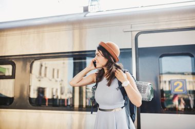 Tema ulaşım ve seyahat. Dişlek gülümseme ayakta portre Genç beyaz kadınla İstasyonu Tren arka plan teknolojisini kullanarak sırt çantası ile tren, telefon el elbise ve şapka yaz aylarında akıllı.