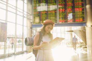 Tema seyahat ve ulaşım. Güzel genç beyaz kadın elbise ve tren istasyonu terminal seyir elektronik çetele tutma telefon, harita kağıt el gezinti içinde ayakta sırt çantası.