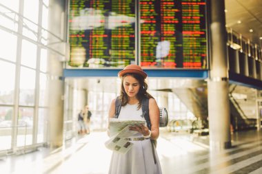 Tema seyahat ve ulaşım. Güzel genç beyaz kadın elbise ve tren istasyonu terminal seyir elektronik çetele tutma telefon, harita kağıt el gezinti içinde ayakta sırt çantası.