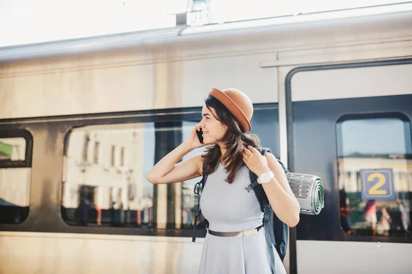 Theme transportation and travel. Portrait young caucasian woman with toothy smile standing train station train background with backpack using technology, smart phone hands in dress and hat summer.