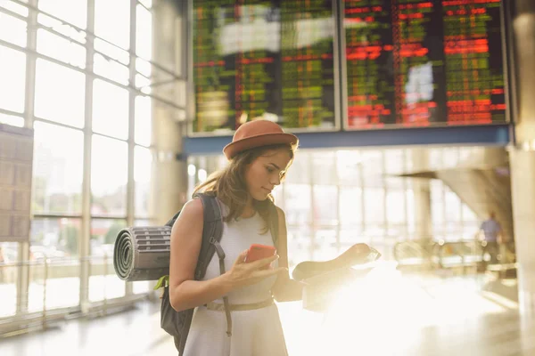 Tema seyahat ve ulaşım. Güzel genç beyaz kadın elbise ve tren istasyonu terminal seyir elektronik çetele tutma telefon, harita kağıt el gezinti içinde ayakta sırt çantası.