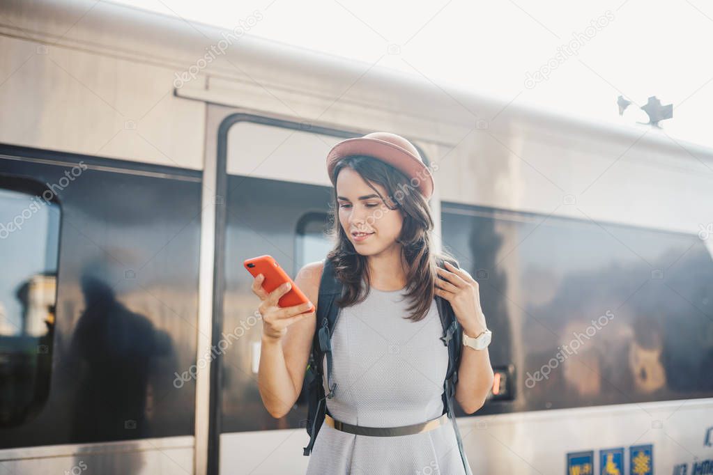 Theme transportation and travel. Portrait young caucasian woman with toothy smile standing train station train background with backpack using technology, smart phone hands in dress and hat summer.