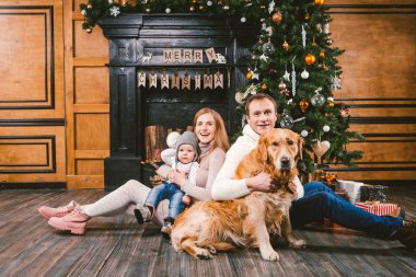 Tema Noel ve yeni yıl aile çevresi. Genç beyaz aile ile 1 yaşındaki çocuk köpek doğurmak Labrador Golden Retriever ahşap zemin oturma odasında şömine Noel ağacı yakınındaki ev üzerinde oturan.