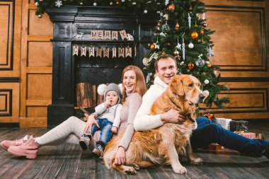 Tema Noel ve yeni yıl aile çevresi. Genç beyaz aile ile 1 yaşındaki çocuk köpek doğurmak Labrador Golden Retriever ahşap zemin oturma odasında şömine Noel ağacı yakınındaki ev üzerinde oturan.