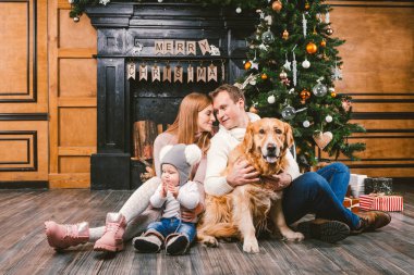 Tema Noel ve yeni yıl aile çevresi. Genç beyaz aile ile 1 yaşındaki çocuk köpek doğurmak Labrador Golden Retriever ahşap zemin oturma odasında şömine Noel ağacı yakınındaki ev üzerinde oturan.