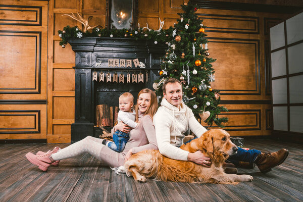 Theme Christmas and New Year family circle. Young Caucasian family with 1 year old child dog breed Labrador Golden Retriever sitting on wooden floor home in living room near fireplace Christmas tree.
