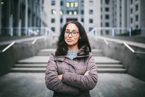 Beautiful young woman of European ethnicity with long brunette hair with grave emotion, wearing glasses and a coat stands against the backdrop of a business center in fall in cloudy cloudy weather.