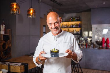 Yakışıklı İtalyan adam kel beyaz üniforma şef onun elinde kazma dişli bir gülümseme ile tatlı bir tabak poz bir restoranda bir sakallı.