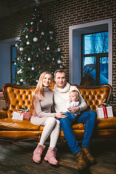 theme new year and Christmas holidays in family atmosphere. Mood celebrate Caucasian young mom dad and son 1 year old sit on a leather brown sofa at home in the living room near the Christmas tree.