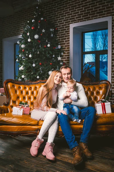 theme new year and Christmas holidays in family atmosphere. Mood celebrate Caucasian young mom dad and son 1 year old sit on a leather brown sofa at home in the living room near the Christmas tree.