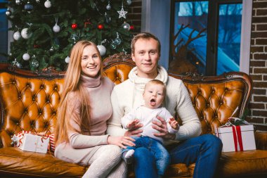 Tema yeni yıl ve Noel tatili aile ortamı içinde. Ruh kutlamak beyaz genç anne baba ve oğlu 1 yaşında oturup Noel ağacı yakınındaki oturma odasında evde deri kahverengi kanepe.