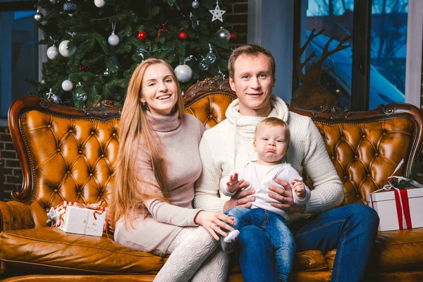 theme new year and Christmas holidays in family atmosphere. Mood celebrate Caucasian young mom dad and son 1 year old sit on a leather brown sofa at home in the living room near the Christmas tree.