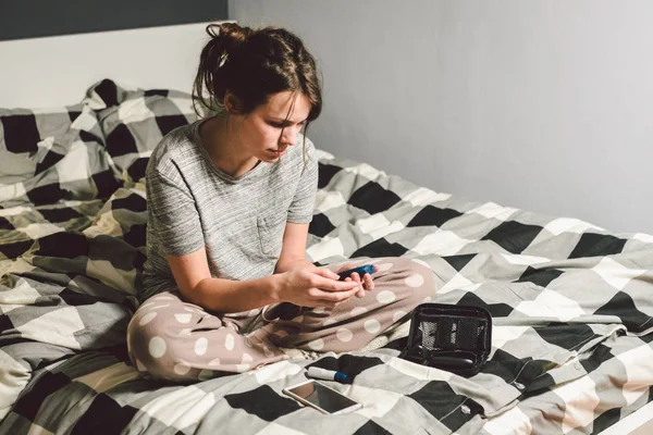 Theme diabetes. young caucasian woman at home in the bedroom on the bed uses the technology of the device for measuring blood glucose, glucometer test strips high sugar level disease.