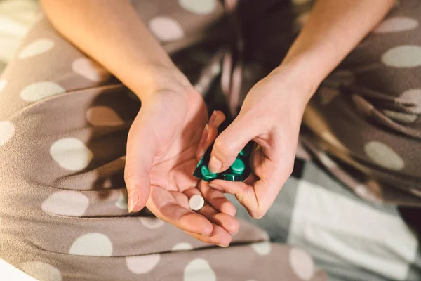 Subject medicine health and pharmaceuticals. Close-up macro young caucasian woman hands pulling out a green blister. Packing two white round pills in home clothes at home on the bed.