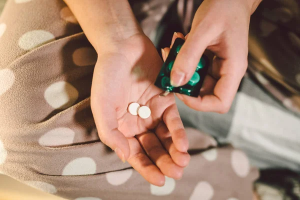 Subject medicine health and pharmaceuticals. Close-up macro young caucasian woman hands pulling out a green blister. Packing two white round pills in home clothes at home on the bed.