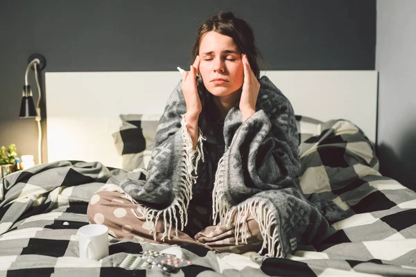 Subject disease and treatment. young Caucasian woman sits home bedroom bed wrapped in body blanket with high fever, fever holds strong headache behind head, uses drop medicine in nose for sinusitis — Stock Photo, Image