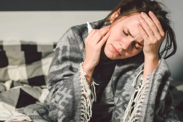 Хвороби та лікування. Молода кавказька жінка сидить вдома у ліжку з ліжком, загорнутим у ковдру з високою температурою, гарячка має сильний головний біль за головою, застосовує ліки від синуситу в носі. — стокове фото