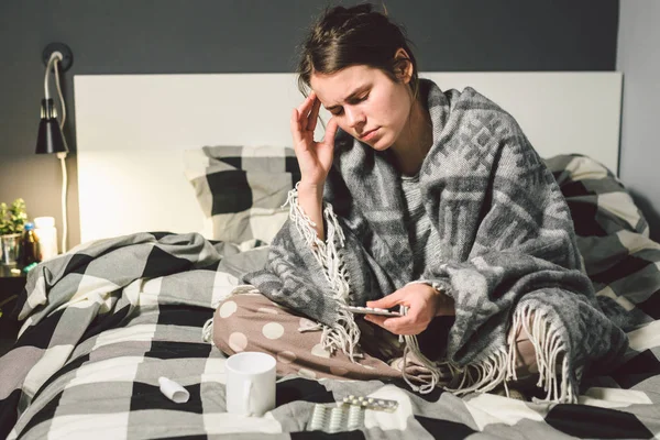 Sujeto enfermedad y tratamiento. joven mujer caucásica se sienta en casa dormitorio cama envuelta en manta corporal con fiebre alta, fiebre sostiene fuerte dolor de cabeza detrás de la cabeza, utiliza medicamentos de gota en la nariz para la sinusitis — Foto de Stock