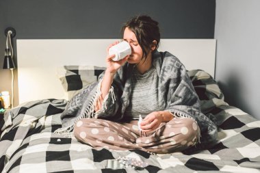 Young beautiful Caucasian woman has a cold, flu with high fever and heat at home in bed. The girl takes headache pills with a glass of water, a bitter pill is crooked. clipart