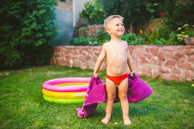 Yaz tatil tema. Kuru bir havlu kırmızı yüzme şort olmak yok, yuvarlak bir şişme renkli havuz yakınındaki çimenlerin üzerinde bir evin arka bahçesinde oynarken küçük 3 yaşındaki beyaz çocuk sarılmış.