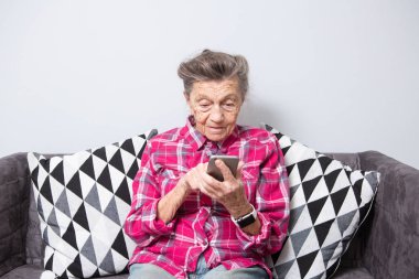 Tema eski kişi teknolojisini kullanır. eski gri saçlı beyaz bir kadın kırışıklıkları oturuyor ellerini, kanepe ve kullandığı cep telefonunda oturma odasında ev ile belgili tanımlık perde, parmak dokunmatik ekran üzerinde görünüyor.