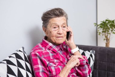 Tema eski kişi teknolojisini kullanır. Arama ve telefonda konuşurken evde oturma odasında kanepede oturan ve bir cep telefonu kullanarak olgun mutlu sevinç gülümseme etkin gri saç beyaz kırışıklıkları bir kadın.