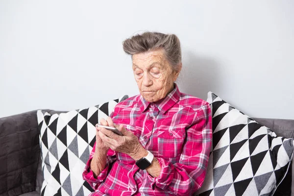 Theme old person uses technology. old gray-haired Caucasian woman with wrinkles sits home in living room on sofa and uses mobile phone in hands, looks the screen, finger on touch screen.