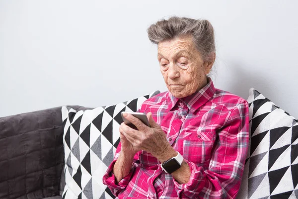 Theme old person uses technology. old gray-haired Caucasian woman with wrinkles sits home in living room on sofa and uses mobile phone in hands, looks the screen, finger on touch screen.