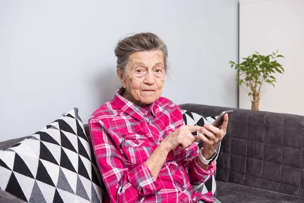 Theme old person uses technology. old gray-haired Caucasian woman with wrinkles sits home in living room on sofa and uses mobile phone in hands, looks the screen, finger on touch screen.