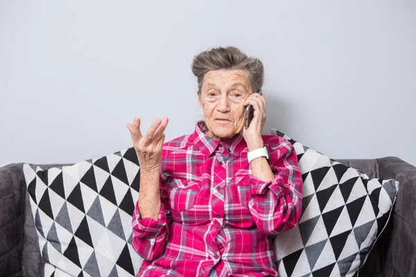 Theme old person uses technology. Mature happy joy smile active gray hair Caucasian wrinkles woman sitting home in living room on the sofa and using a mobile phone, calling and talking on the phone.