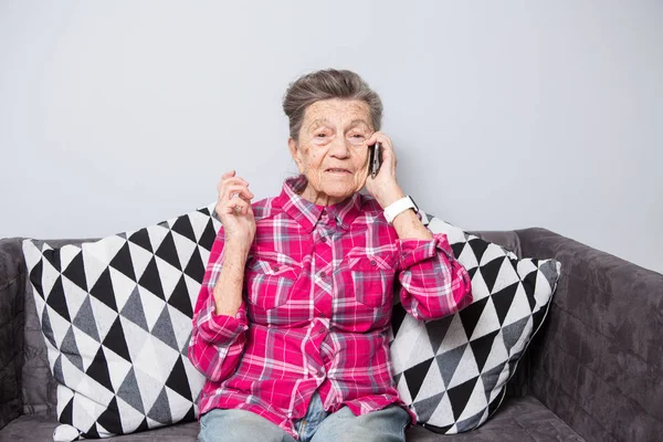 Theme old person uses technology. Mature happy joy smile active gray hair Caucasian wrinkles woman sitting home in living room on the sofa and using a mobile phone, calling and talking on the phone.