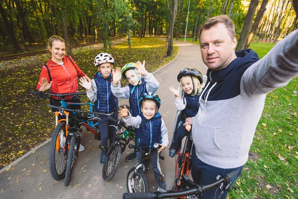 The theme family sports outdoor recreation. large family Caucasian 6 people mom dad and 4 children three brothers and sister ride bicycles in park on bicycle path. Father holds camera makes photo.