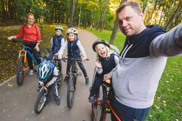 The theme family sports outdoor recreation. large family Caucasian 6 people mom dad and 4 children three brothers and sister ride bicycles in park on bicycle path. Father holds camera makes photo.