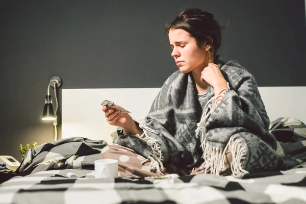 A young Caucasian woman sits on a bed in a bedroom at home holding many packs of blister tablets and medicines in her hands. Cold fever and temperature. Theme cold and illness.