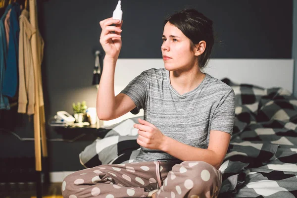 The theme is seasonal cold, runny nose flu virus infection. Young caucasian woman at home bedroom bed uses spray drops of medicine in her nose for snot. Allergic renitis and sinusitis.