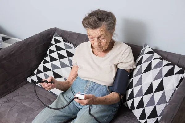 The topic is very old person and health problems. A senior Caucasian woman, 90 years old, with wrinkles and gray hair, sits home on sofa and uses blood pressure monitor to measure blood pressure.