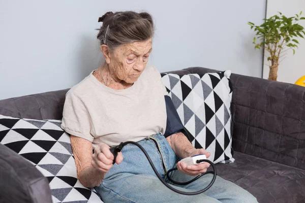话题是非常老的人和健康问题 0岁的白种人老年女性 有皱纹和灰白的头发 坐在家里的沙发上 用血压监测仪测量血压 — 图库照片