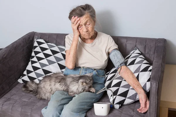 The topic is very old person and health problems. A senior Caucasian woman, 90 years old, with wrinkles and gray hair, sits home on sofa with pet cat and uses tonometer. High blood pressure headache.