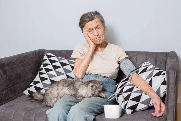 The topic is very old person and health problems. A senior Caucasian woman, 90 years old, with wrinkles and gray hair, sits home on sofa with pet cat and uses tonometer. High blood pressure headache.