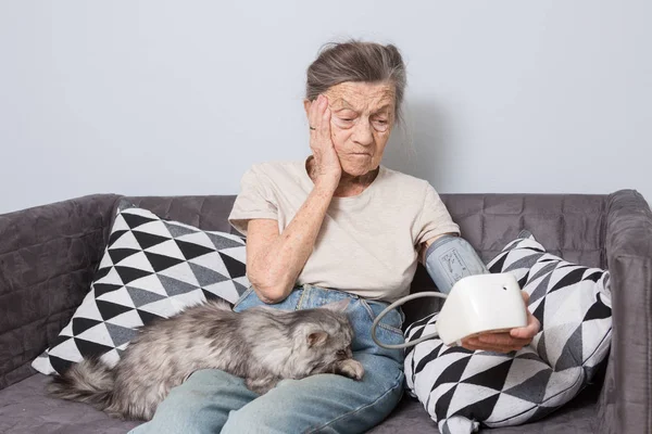 话题是非常老的人和健康问题 一位90岁的高级白人妇女 带着皱纹和灰白的头发 和宠物猫坐在家里的沙发上 使用眼压计 高血压头痛 — 图库照片