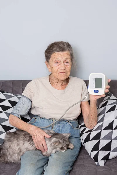 The topic very old person and monitor health. senior Caucasian woman, 90 years old, with wrinkles and gray hair, sits home on a sofa with a pet cat and uses a tonometer. Good mood healthy pensioner.