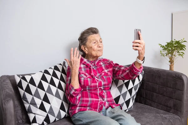 A very old senior Caucasian grandmother with gray hair and deep wrinkles sits home on couch in jeans and red checkered shirt and uses her smart phone on outstretched arm front camera for a video call.