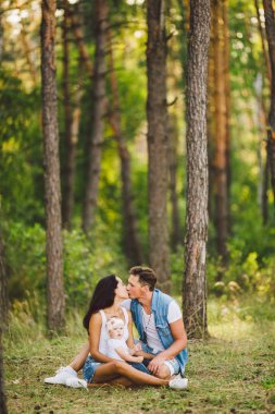 Tema aile açık rekreasyon orman park alanında. Genç güzel şık beyaz aile küçük çocuk kızı bir yaşında ile oturup iğne yapraklı orman çim üzerinde yaz gün batımında.