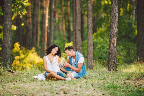森林公園地区テーマ家族野外レクリエーション 歳の小さい子娘と若い美しいスタイリッシュな白人家族日没で夏に針葉樹林の草の上に座る — ストック写真