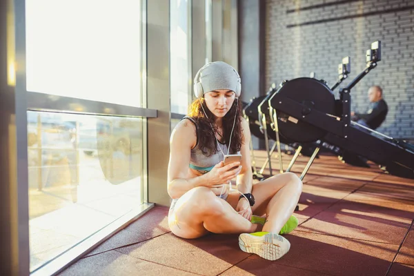 Theme sports, health and technology. beautiful sexy Caucasian woman sportswoman in gray sportswear and hat sits by window with sun setting uses smartphone listen music in large headphones on head.