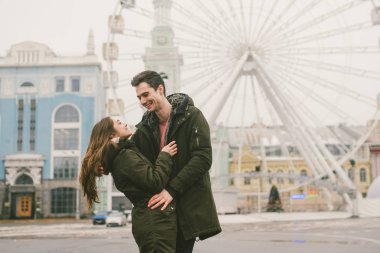 Tema aşk ve tatil Sevgililer günü. Beyaz heteroseksüel aşıklar çifti birlikte kasvetli hava kucaklama çerçevede dönme dolap kasaba Meydanı'nda kış. Adam yavaşça kız hugs.